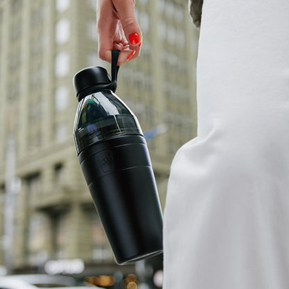 Person holding a black KeepCup water bottle by the carry hang with a blurred cityscape background