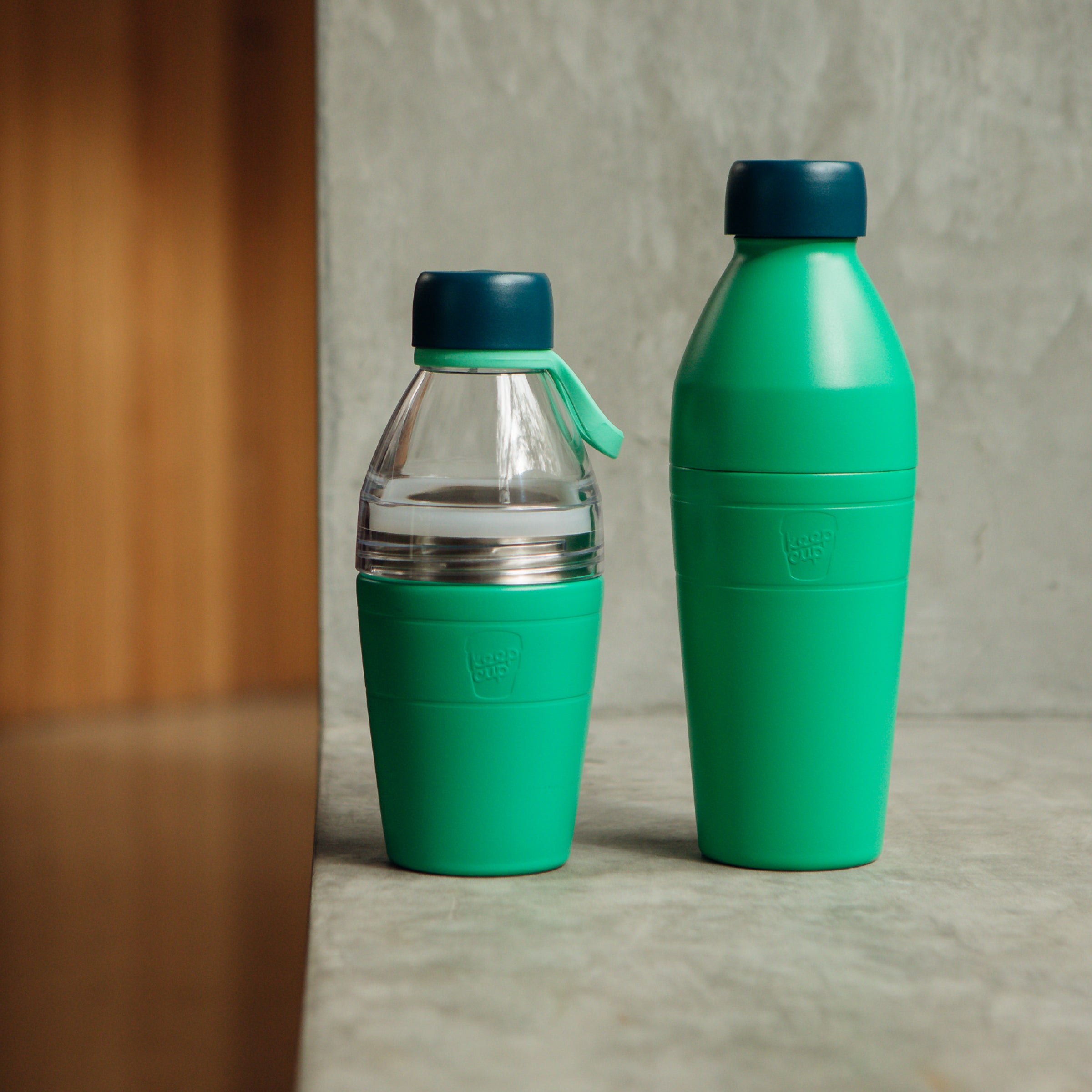 Two green KeepCup water bottles with blue lids on a gray surface.