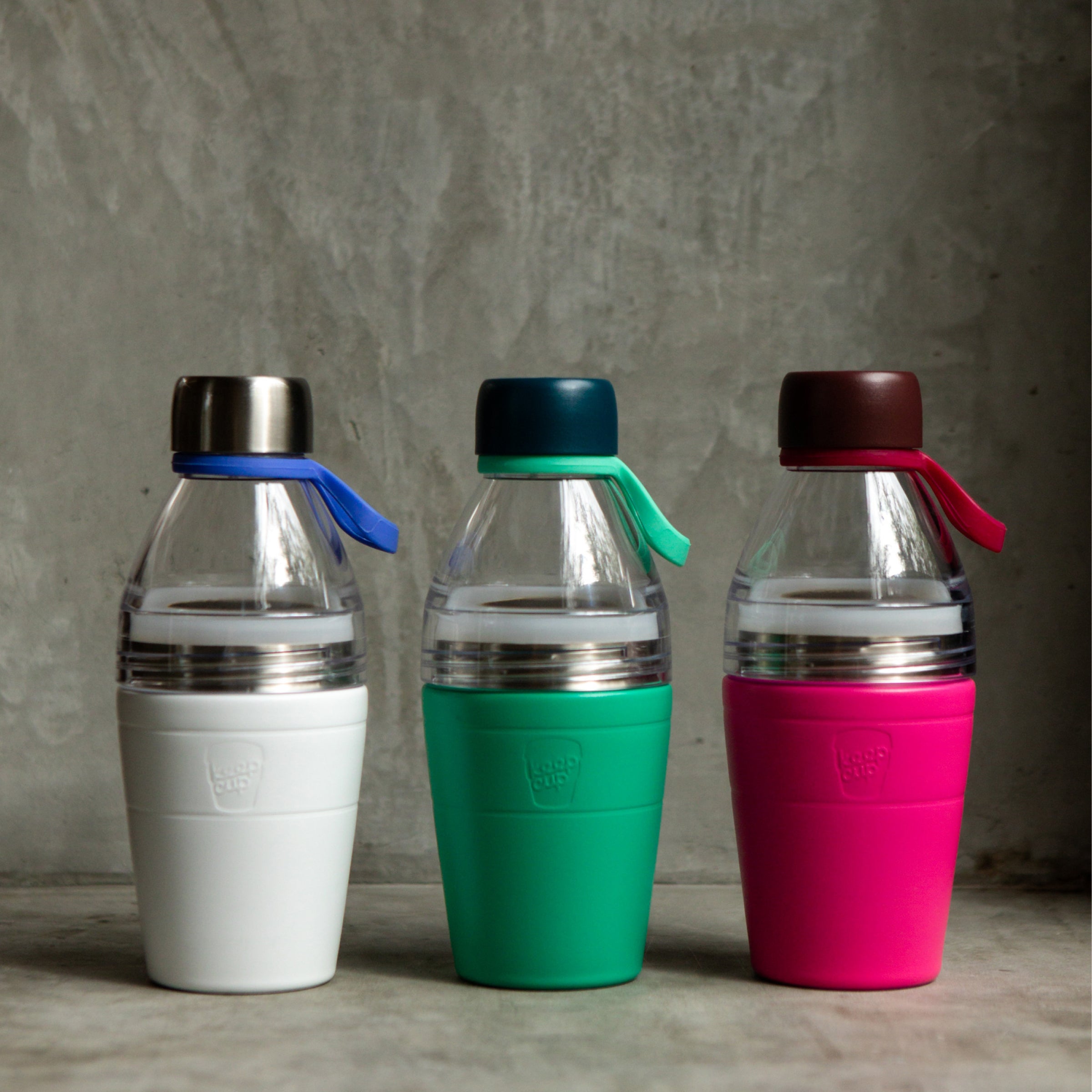 Three colorful KeepCup water bottles, one white, green and red with different colored lids on a gray surface.