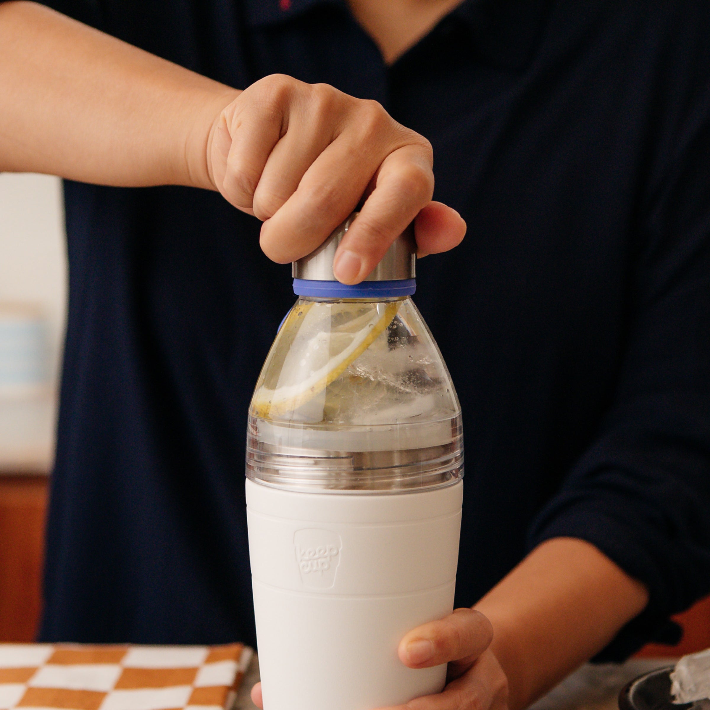 Person opening a white KeepCup water bottle with a lemon slice inside, kitchen setting with blurred background