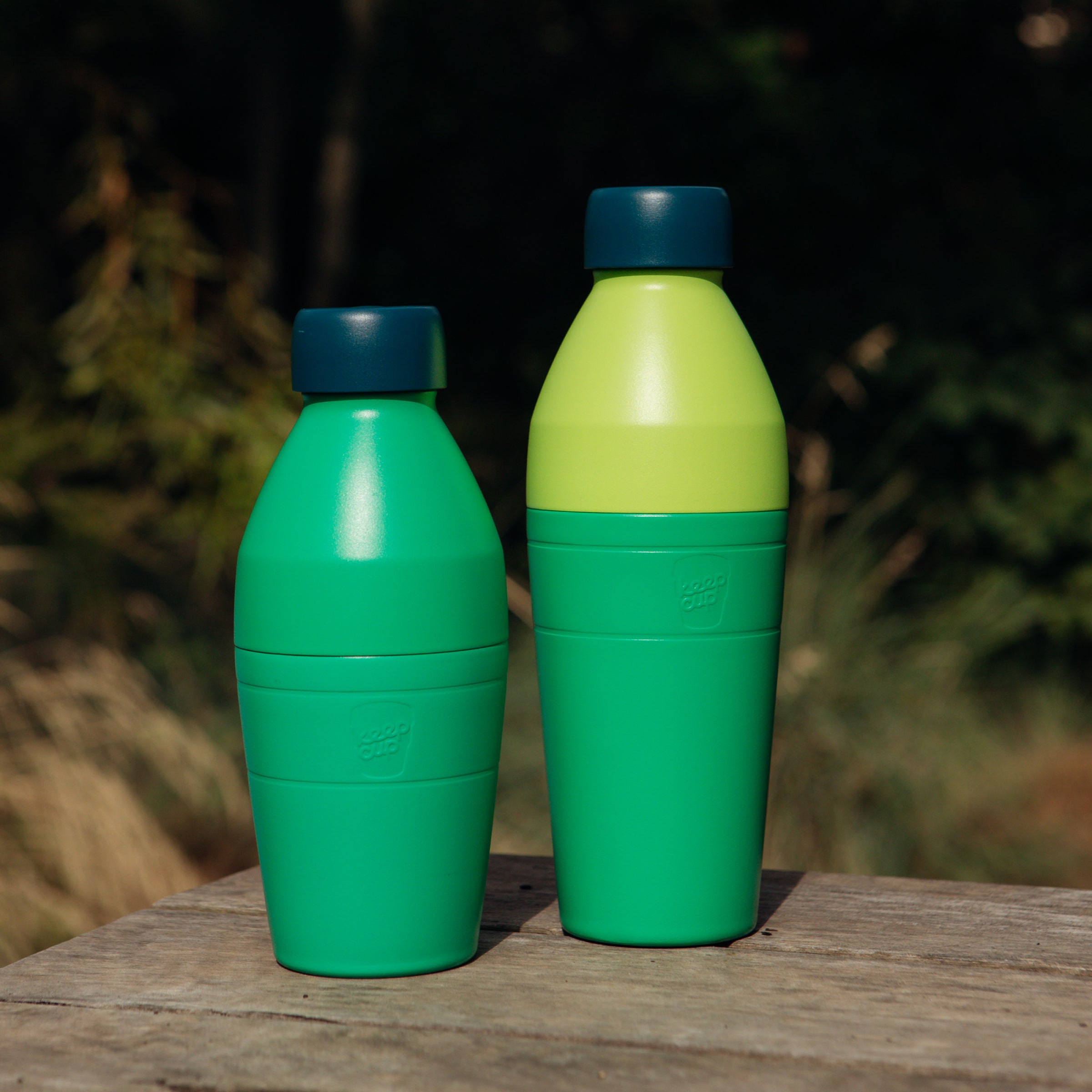 Two KeepCup green bottles with blue lids on a wooden surface outdoors.