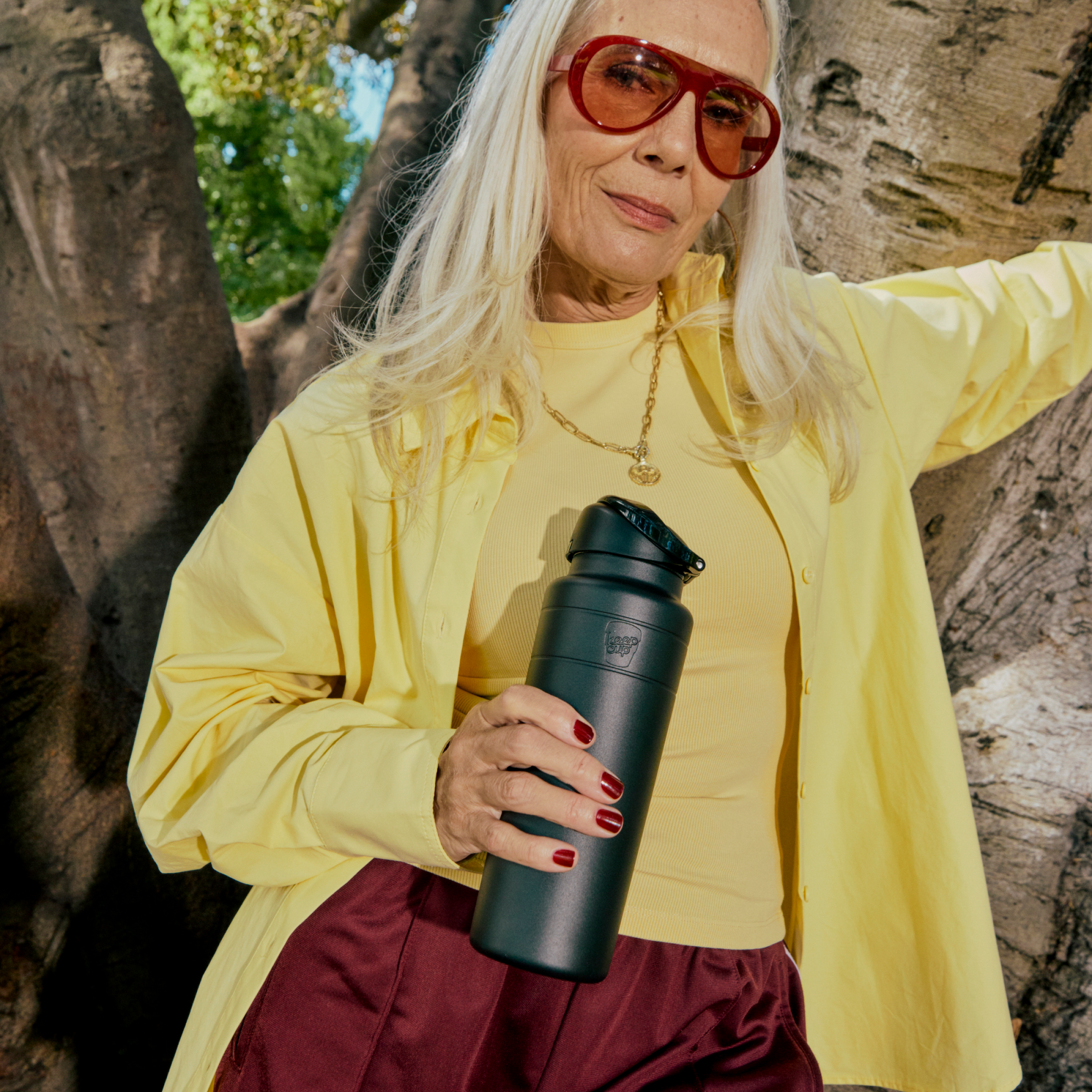 A woman with long white hair wearing red sunglasses and a yellow outfit holds a KeepCup Ora Bottle 24oz in Black while leaning against a tree trunk outdoors.