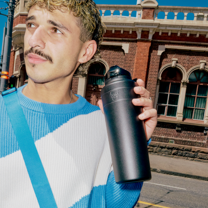 A man with curly hair and a mustache wearing a blue and white striped sweater holds a KeepCup Ora Bottle 32oz in Black while standing on an urban street with a historic brick building in the background.