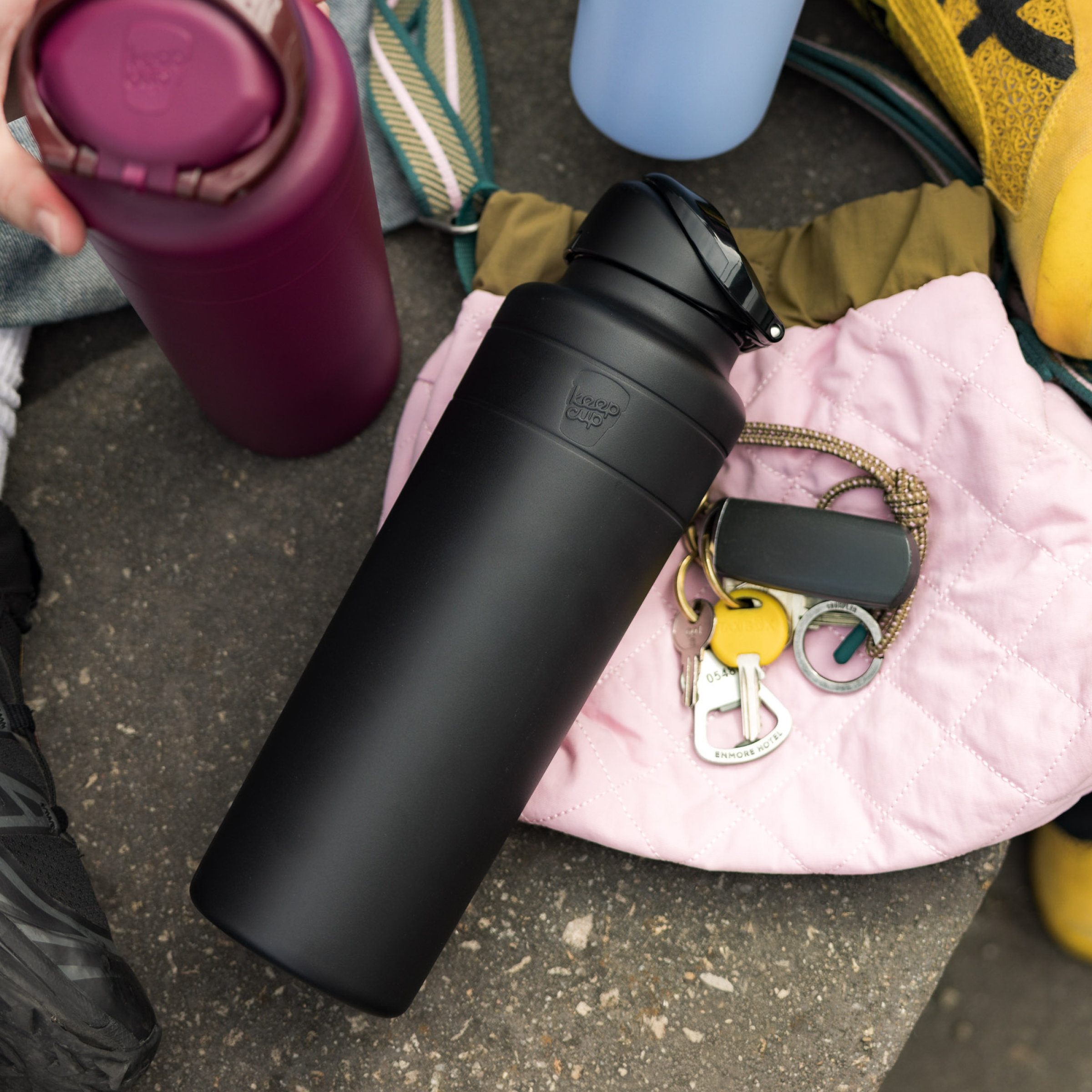 KeepCup Ora Bottle 32oz in Black lying on a pink quilted jacket beside keys and a car remote, with a hand holding a burgundy KeepCup nearby, surrounded by everyday items like sneakers and bags on a concrete surface.