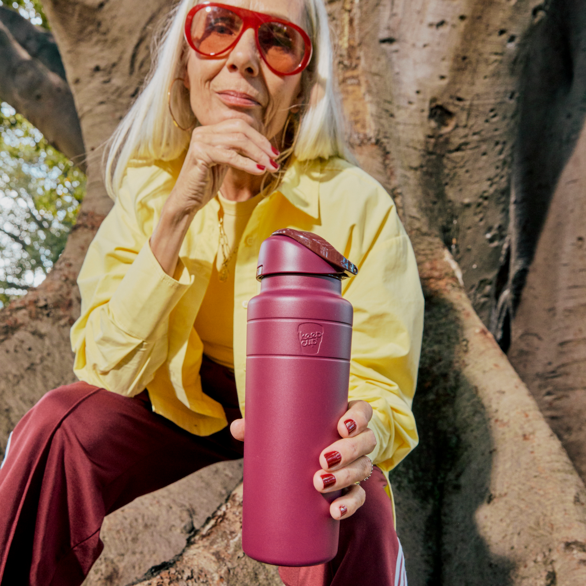 A woman with silver hair and red sunglasses sits on a large tree trunk, holding out a KeepCup Ora Bottle 24oz in Mulberry color toward the camera, wearing a yellow button-up shirt and burgundy pants with matching red nail polish.
