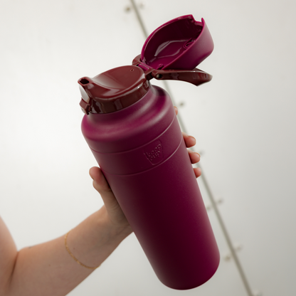 A hand holds the KeepCup Ora Bottle 32oz in Mulberry at an angle, showcasing the deep purple matte finish and open flip-top lid against a neutral background.
