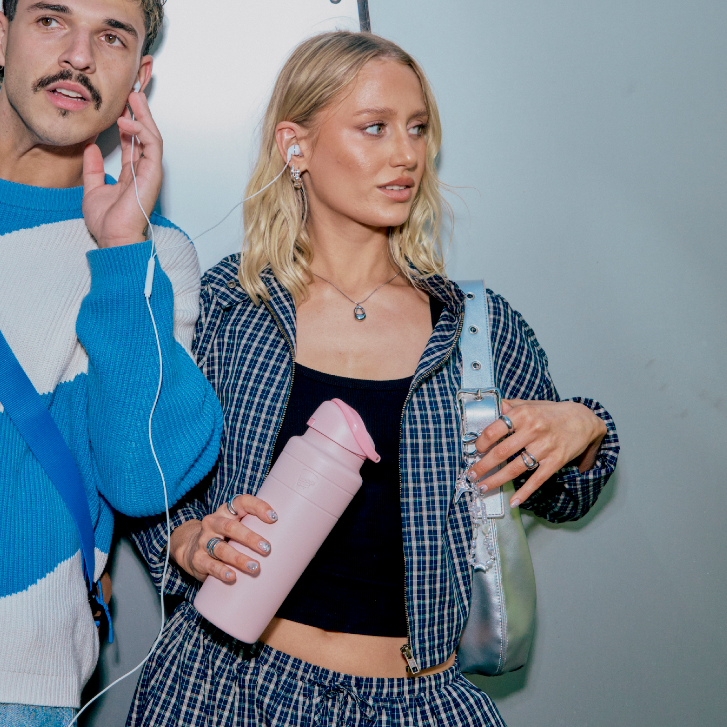 A young woman with blonde wavy hair holds the Ora Bottle 24oz in Pashmak Pink while wearing a blue plaid jacket over a black crop top, standing next to a man in a blue and white sweater sharing earbuds with her.