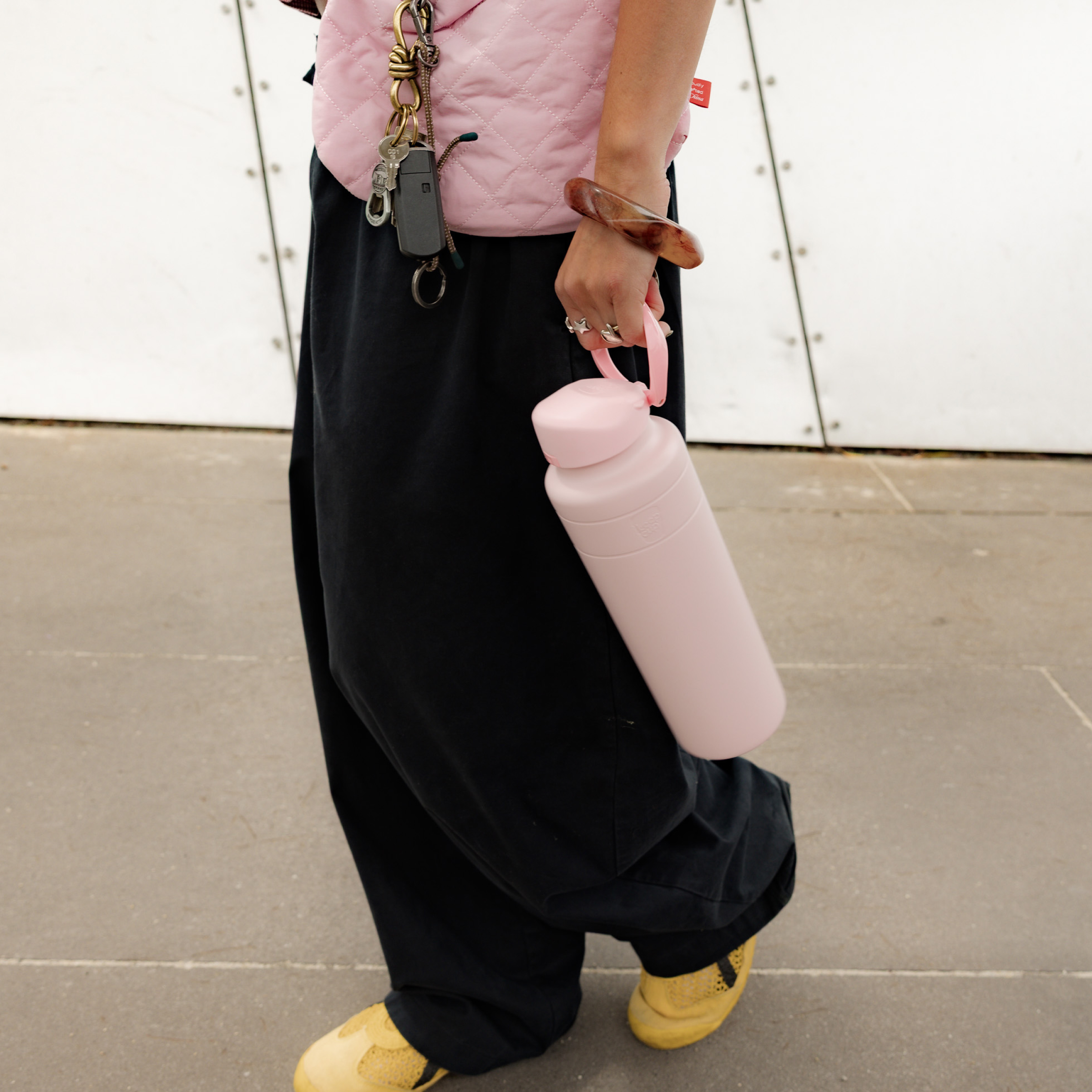 Person walking outdoors holding a KeepCup Ora Bottle 32oz in Pashmak Pink by the carry strap, styled with a matching pink quilted vest, black wide-leg pants, and yellow sneakers.