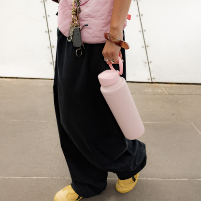 Person walking outdoors holding a KeepCup Ora Bottle 32oz in Pashmak Pink by the carry strap, styled with a matching pink quilted vest, black wide-leg pants, and yellow sneakers.