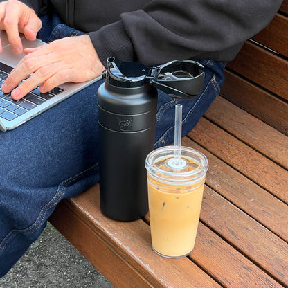 A person sits on a wooden bench working on a laptop, with the KeepCup Ora Bottle 24oz in Black featuring an open flip-top lid resting beside them, along with an iced coffee in a clear reusable cup with a straw.
