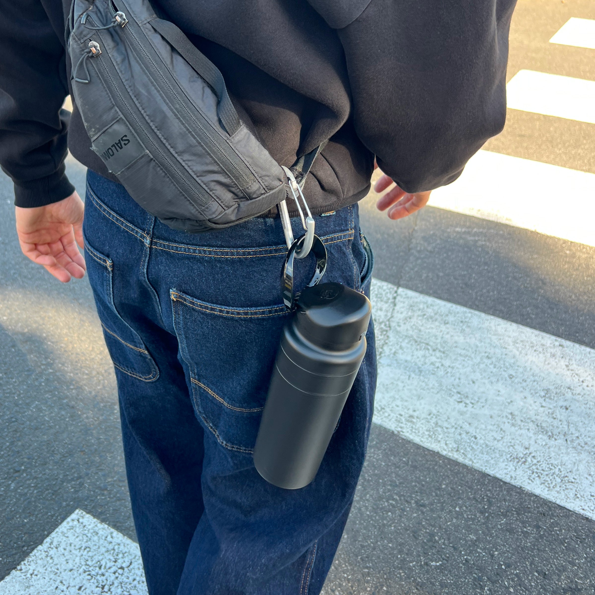Person walking near a crosswalk wearing dark jeans and a black hoodie with a Salomon waist bag, the KeepCup Ora Bottle 24oz in Black clipped to their belt loop via a carabiner.