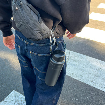 Person walking near a crosswalk wearing dark jeans and a black hoodie with a Salomon waist bag, the KeepCup Ora Bottle 24oz in Black clipped to their belt loop via a carabiner.