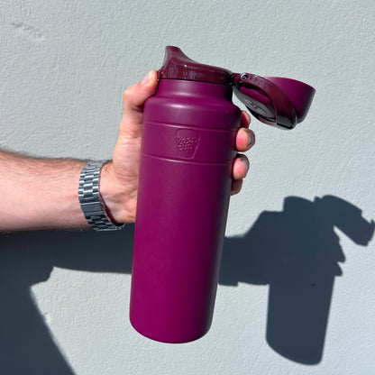 A hand holding the Ora Bottle 32oz in Mulberry with the flip-top lid open, shown against a white textured wall with the bottle's shadow visible in bright sunlight.