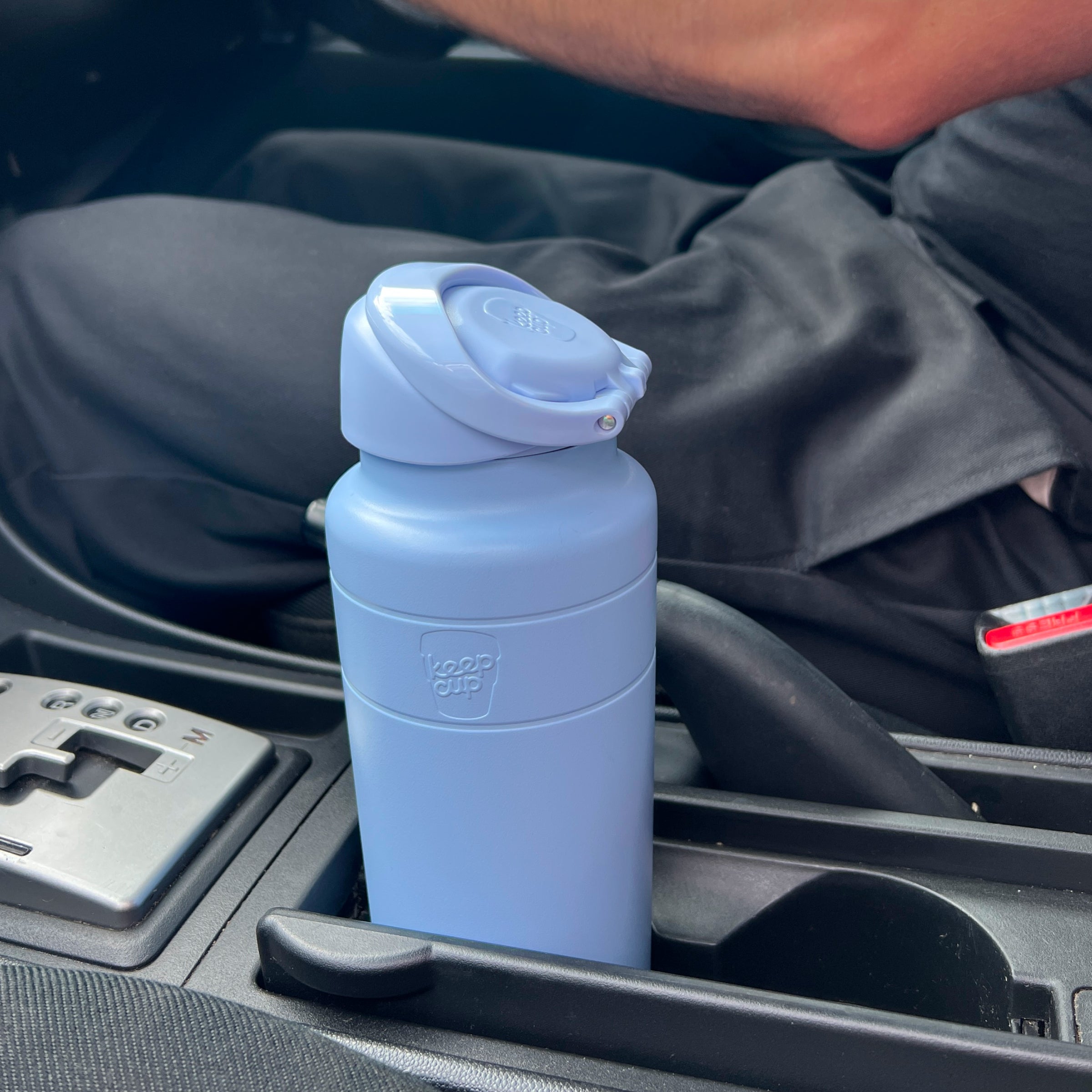 KeepCup Ora Bottle 24oz in Orchid, a matte periwinkle blue insulated water bottle with flip-top lid, sitting in a car cup holder next to the center console and gear shift.