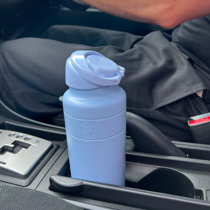 KeepCup Ora Bottle 24oz in Orchid, a matte periwinkle blue insulated water bottle with flip-top lid, sitting in a car cup holder next to the center console and gear shift.