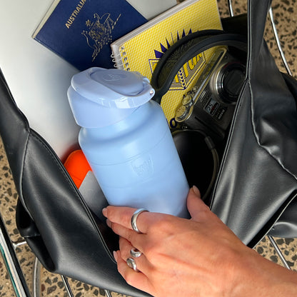 A hand with silver rings places a KeepCup Ora Bottle 24oz in Orchid into a black leather tote bag alongside travel essentials including an Australian passport, yellow notebook, Panasonic Lumix camera, and sunscreen.