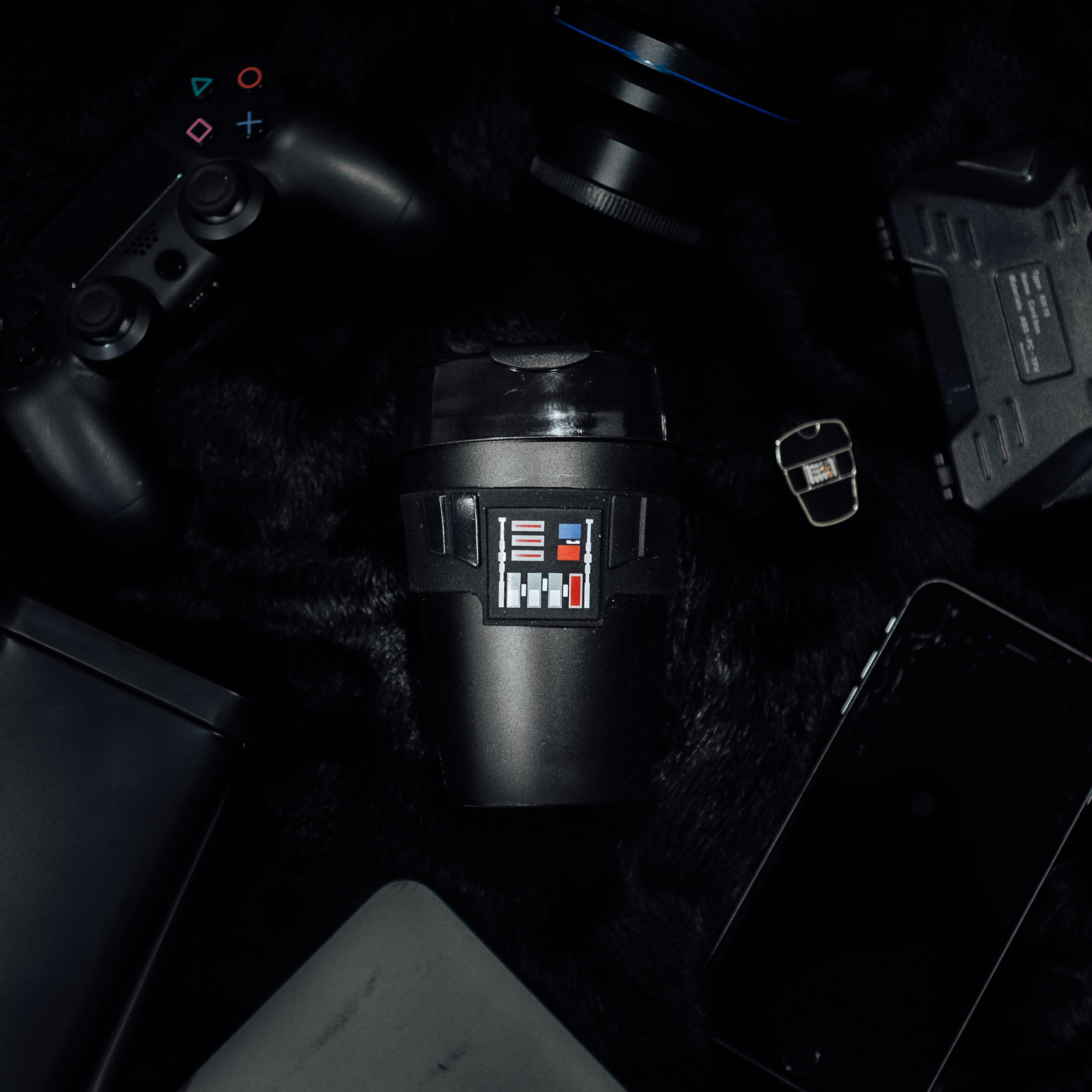 Darth Vader Original M 12oz KeepCup featuring the iconic black design with chest panel graphics, shown from above on a dark furry surface surrounded by tech gadgets including a PlayStation controller, camera lens, smartphone, and tablet.