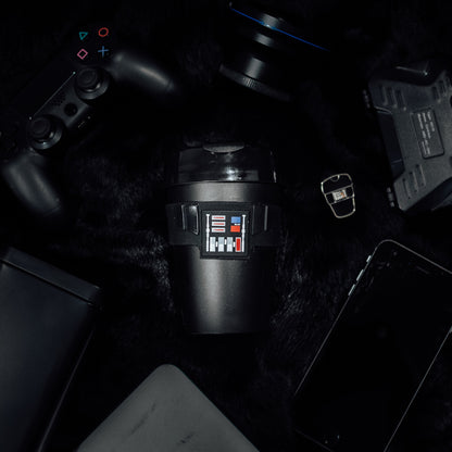 Darth Vader Original M 12oz KeepCup featuring the iconic black design with chest panel graphics, shown from above on a dark furry surface surrounded by tech gadgets including a PlayStation controller, camera lens, smartphone, and tablet.