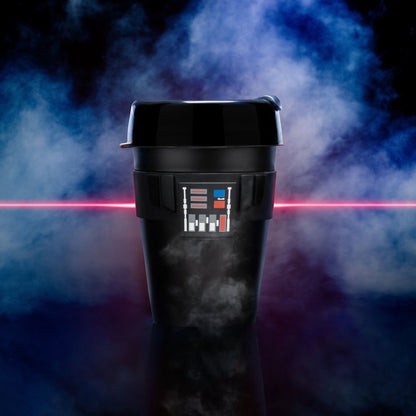 Darth Vader Original M 12oz black reusable coffee cup featuring the iconic chest panel design, displayed against a dramatic smoky blue and purple background with a red lightsaber beam of light crossing horizontally behind it.
